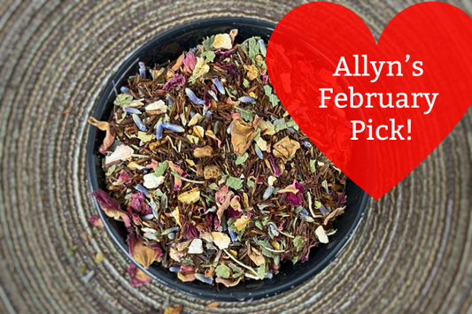 Bowl of dried herbs with a red heart-shaped card reading 'Allyn's February Pick!' on a textured surface.
