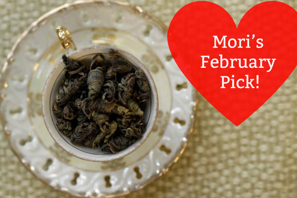Tea leaves in a white teacup with a red heart-shaped label reading 'Mori's February Pick!' on a textured beige background.