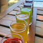 Row of rainbow colored teas in small glasses on a wooden table outdoors.