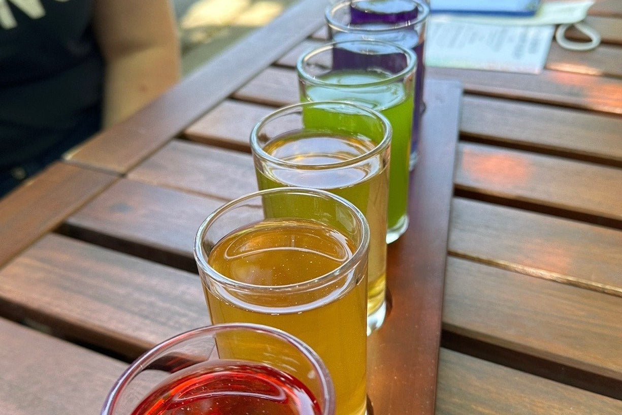 Row of rainbow colored teas in small glasses on a wooden table outdoors.